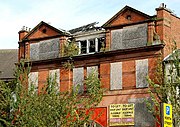 Upper floors of a red-brick building with boarded windows. The top floor features twin porticoes, with a stone porthole below the apex of each roof.