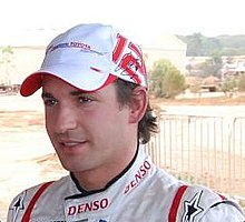 Head and shoulders of a man in his twenties with dark brown hair and eyes. He is wearing a red-and-white baseball cap which displays the Panasonic Toyota Racing logo at the front, and the number 12, with his signature superimposed upon it, on the left side. He is also wearing white racing overalls with red stripes, upon the collar of which the Denso logo is embroidered.