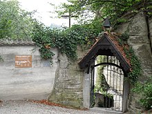 In der Mauer befindet sich ein Gittertor. Es ist mit einem etwa 50 cm vorstehenden Giebeldach, belegt mit Biberschwanzziegeln, bekrönt. Die Mauer ist noch etwa 30 cm höher als der Dachgiebel.