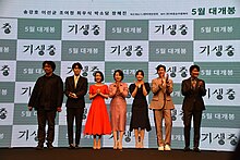 Photo de quatre hommes et trois femmes sur une scène devant un écran affichant des logos en coréen. Ils regardent vers le public, la plupart en souriant et en applaudissant.