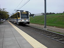 A white and yellow light rail train approaches an empty station platform with overhead lines visible above.