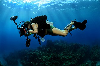 A scuba diver swims over a reef with a large still camera in an underwater housing with dome port and electronic strobes.