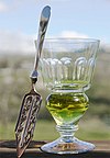 A reservoir glass filled with a naturally colored verte next to an absinthe spoon.