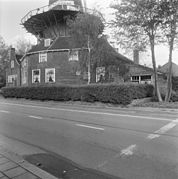 Photographie en noir et blanc d'une maison et d'une remise couronnés d'une galerie et longées par une route.