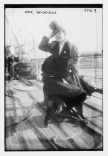 Black and white photograph of a middle-aged woman standing on a dock or the deck of a ship. Her legs are crossed and she is holding her hat.