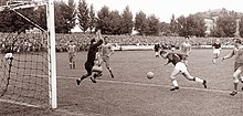 Черно-белое фото, на котором футболист отбивает мяч ногой в сторону ворот, в то время как вратарь соперника пытается сделать сейв.