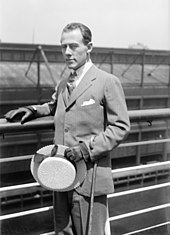 Photographie en noir et blanc d'un homme en costume tenant un chapeau dans sa main gauche.