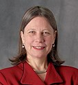 A color photograph of a smiling woman with blue eyes wearing a red shirt, a necklace, and pearl earrings, all in front of a grey background