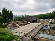 Fotografie archeologického naleziště – je vidět základy řady rozlehlých budov, dřevěné chodníky mezi nimi, informační cedule, domek uprostřed areálu. Vše na mírném svahu, obklopeno hustým lesem.