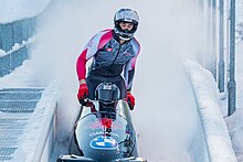 Colour photograph of Mike Evelyn standing up in a black BMW bobsleigh with the Canadian maple leaf on the front.