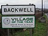 Дорожные знаки с черной надписью Backwell на белом фоне, а под ним еще один знак с надписью «Деревня года Юго-Запад» и региональный победитель Уэльса 1997 года.