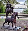 Seitenansicht der Bronzeskulptur mit Hund, Affe, Kaninchen und Ratte. Der Affe hat den Mund weit geöffnet, als würde er schreien. Am Boden vor der Skulptur eine Steinplatte mit Gravur sowie Beetpflanzen, im Hintergrund eine Bank, Bäume und die Außenalster.