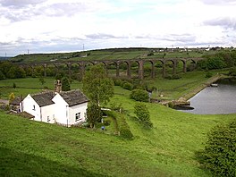 A white cottage in the foreground, a dam head and water in the middle ground, and a railway viaduct in the background