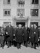 Photo en noir et blanc d’un groupe d'individus marchant dos à un bâtiment officiel ; au centre, un homme aux cheveux gris-blancs et à la moustache grise tient un chapeau dans sa main droite et une canne sous son bras gauche