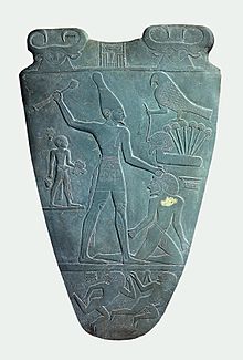 The Narmer Palette. The face of a woman with the horns and ears of a cow, representing Bat or Hathor, appears twice at the top of the palette, and a falcon representing Horus appears to the right of the palette. Narmer Palette smiting side.jpg