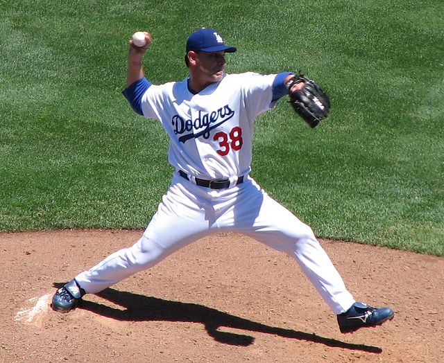 A man wearing a white baseball uniform with blue script across the chest and a blue baseball cap throwing a baseball with his right hand from a dirt mound on a grass field