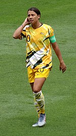 Photographie de l'attaquante australienne Sam Kerr sur un terrain de football avec le maillot de l'équipe nationale et le brassard de capitaine.