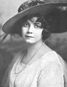 A young white woman with dark hair and eyes, wearing a wide-brimmed hat, a strand of pearls, and a white dress or blouse with a square neckline