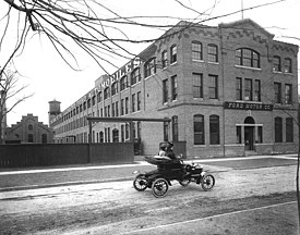 A black and white photograph of a brick building, with a sign on the front saying "Ford Motor Co.', in front of which is two people in a car