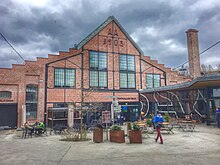 Foto eines Gebäudes mit roter Ziegelfassade mit der Aufschrift. Vor dem Gebäude befinden sich Tische und Stühle.