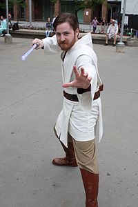 Homme à barbe portant une tunique blanche et une réplique de sabre laser.