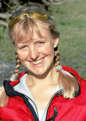 Daniela Jasper ist im porträt frontal lächelnd in die Kamera schauend abgebildet. Sie hat blondes zu zwei Zöpfen geflochtenes Haar, welche auf Schulterhöhe enden. Sie trägt einen roten sportlich wirkenden Pullover mit Reißverschluss, der aufgezurrt ist. Im Hintergrund ist eine Wiese oder grüne Landschaft auszumachen.
