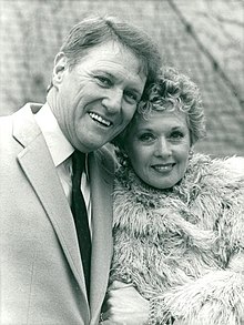 A black-and-white photo of two middle-aged Caucasians; a man and woman. The man with short, light-colored hair, wears a light-colored suit with a dark tie, and is smiling towards the camera. The woman, who also has short, light-colored hair, wears a fur coat, and is also looking towards the camera with a smile.