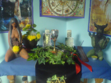 Wicca-Altar mit Statuen von Göttin und Gott, einem Kessel, Kelch, Blumen und Kerzen. Im Hintergrund ist das Rad des Jahres abgebildet.