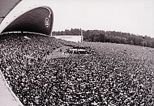 A large meeting of people assembled in a park in front of an arch-like structure, on which the Columns of Gediminas, a XIV-century Lithuanian symbol, appear