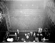 Φωτογραφία παρμένη από το ταβάνι ενός βιομηχανικού δωματίου με σχήμα τετραγώνου. Οι τσιμεντένιοι τοίχοι έχουν μεταλλικές σκάλες. Μία ντουζίνα ανθρώπων απασχολούνται με μερικές δραστηριότητες στο πάτωμα