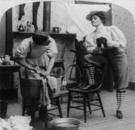 Photo d'une femme portant des knickerbockers et des chaussettes de genou (tenue traditionnelle masculine) et fumant une cigarette, surveillant un homme qui semble porter une robe et un tablier, occupé à la lessive.
