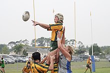 A female player in yellow and green kit and wearing a white scrum cap, jumps to collect a ball while supported by teammates.