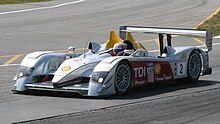 A front three-quarters view of a gloss silver Audi R10 TDI in motion with the number 2 and the words "TDI Power" emblazoned on the side. The driver, Rinaldo Capello, has just begun to turn right.