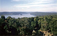 Una vista a través de la selva hacia un lago rodeado de colinas boscosas bajas. Dos pequeñas islas se encuentran cerca de la orilla opuesta. Hay un pequeño claro en el primer plano inmediato. En el lado izquierdo de la orilla opuesta se encuentra una amplia área herbosa frente a las colinas boscosas.