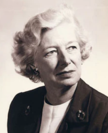 An older white woman with wavy set white hair, wearing a dark suit and white blouse
