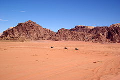 Eine Panorama-Aufnahme mit einer großen rotsandigen Ebene im Vordergrund, auf der, klein in der Bildmitte zu sehen, drei Jeeps in die rechte Richtung fahren. Im Hintergrund erheben sich schroffe Bergfelsen, ebenfalls rötlich. Der Himmel drüber ist blau und wolkenlos.