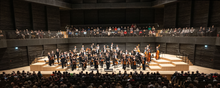 Ein Sinfonieorchester stehend vor Publikum in dunklem Saal