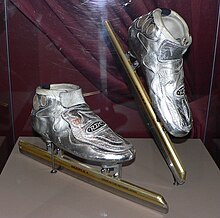 Металлические серебряные коньки с золотыми лезвиями в стеклянной витрине с немного приподнятым правым коньком. За коньками бордовая занавеска. Лезвия намного длиннее, чем сам ботинок коньков.