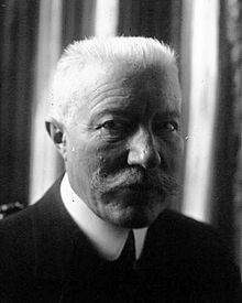 A black and white photo of an older man. He has short white hair and a mustache.