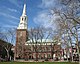 Ground-level side view of a brown brick church with a large, white, tapering spire.