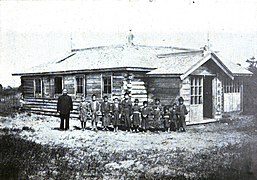 Photo d'un bâtiment en bois de plain-pied, devant lequel posent un groupe d'enfants et leur professeur.