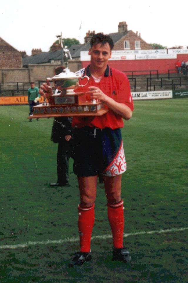 A man standing on a grass football pitch, wearing a red shirt, black shorts and red socks, and holding a trophy.