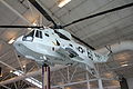 Photographie couleur d'une réplique de l'hélicoptère exposée dans un hangar avec une capsule accrochée