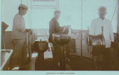 In this picture, on the left side of the picture a sailor is standing on the deck of a metal ship and facing to the right with has his hands on the steering wheel of the ship. In the center of the picture Burton Holmes, in a black suit, is standing and facing to the right with his head turned toward the camera. In front of him is something that looks like a bird bath which might possibly be a big compass. On the right side of the picture, a sailor is standing and facing the camera. In front of him is a pedestal with two handles on it. In the background is the railing of the side of the ship. Beyond that is fog.