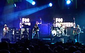 Four men performing on a stage in front of a crowd; three are standing at the front of the stage holding guitars, while one is sitting behind a drum set. Audio equipment, microphone stands, and lighting can also be seen on the stage and in the background.