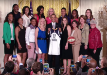 Barack Obama holding up a jersey, smiling, surrounded by the Indiana Fever