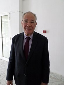 Colour photograph of Mokhtar Trifi aged 78. he is wearing a full suit with a red tie. he is smilling