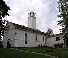 Seitliche Farbfotografie einer weißen Kirche mit einem eckigen Turm in der Mitte. An der linken Mauer ist eine große Nische eingearbeitet und rechts führen Säulenbögen zum Innenhof. Am Kirchenschiff befinden sich schmale Fenster.