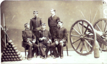 A photograph depicting a group of 5 uniformed men posed between a pyramid of artillery shells on the left and a wheeled field artillery piece on the right