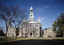 A three-story old stone building with four small spires and a central bell tower, with trees and grass in the foreground.